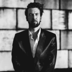 Maurits Chabot staat voor een muur van betonblokken. Hij draagt een pak met een overhemd, zijn handen over elkaar gekruist, zijn nagels zwartgelakt.