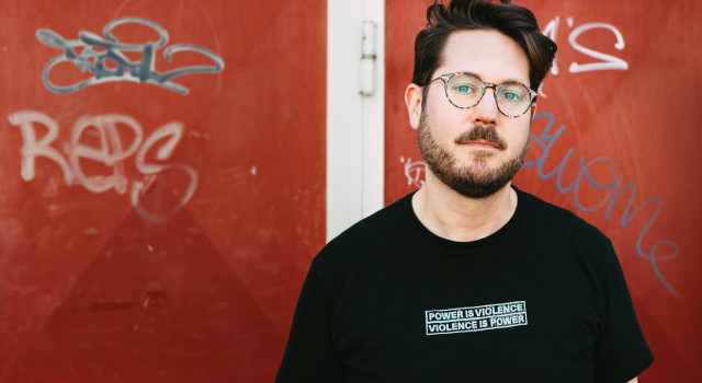 Gijs Wilbrink staat voor een oranje muur met graffiti tags en draagt een bril en een zwart shirt met 'power is violence, violence is power'