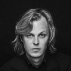 Een zwart-wit portretfoto van Jonah Falke. Hij heeft lang, blond haar, en kijkt recht de camera in.