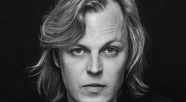 Een zwart-wit portretfoto van Jonah Falke. Hij heeft lang, blond haar, en kijkt recht de camera in.