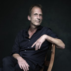 Peter Middendorp zit zijwaarts in een stoel, hij kijkt met een opgetrokken wenkbrauw richting de camera.