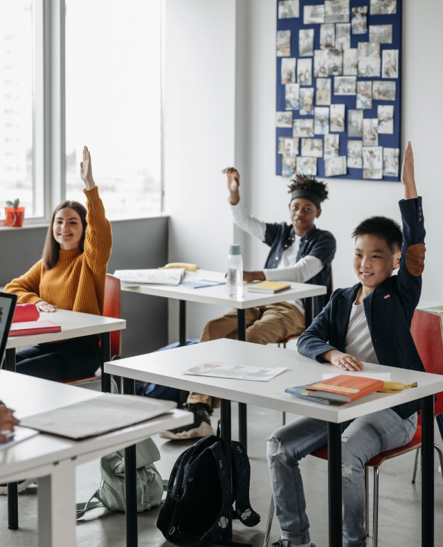 Afbeelding van 4 kinderen aan tafels in een schoolklas met een juf voor de klas