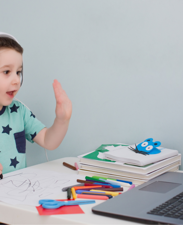 Foto van aan tafel zittend jongetje van ongeveer 5 jaar oud met een koptelefoon op die naar een laptop kijkt en zijn hand opsteekt