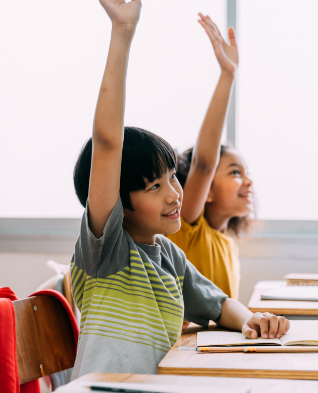 Foto van 2 kinderen zittend aan tafeltjes met hun rechterhand omhoog